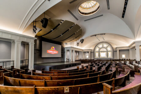A spacious, empty auditorium with curved wooden pews, a large screen displaying 