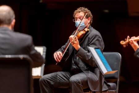 A masked violinist in a black suit performs on stage with other musicians. He sits on a chair, focusing on his music. Sheets of music are visible on the stand in front of him, and the concert hall seatings are dimly lit in the background.
