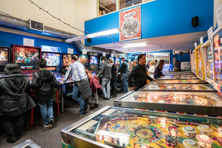 A group of people enjoy playing various pinball machines in a colorful arcade. The room is lively with games and vibrant lights. Walls are painted blue, and the ceiling has string lights. The atmosphere is energetic and nostalgic.