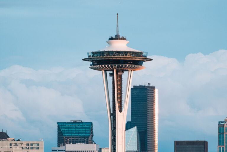 The image shows the Space Needle in Seattle, a tall observation tower with a futuristic design. Surrounding the needle are modern skyscrapers under a clear blue sky with some scattered clouds.