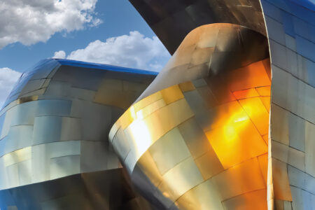 A close-up view of a futuristic building with reflective, metallic surfaces in gold, bronze, and silver tones. The curving structure is set against a blue sky with scattered fluffy clouds, creating an abstract, dynamic appearance.