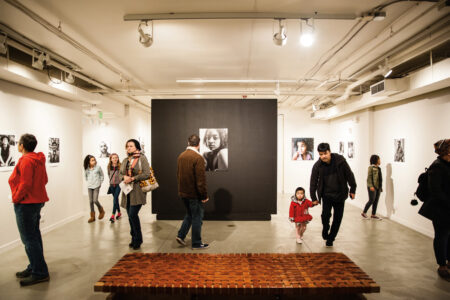 People walk and observe artwork in a spacious gallery. Black and white portraits line the walls, and a large photo is displayed on a central black panel. A wooden bench is in the foreground. The atmosphere is relaxed and engaged.