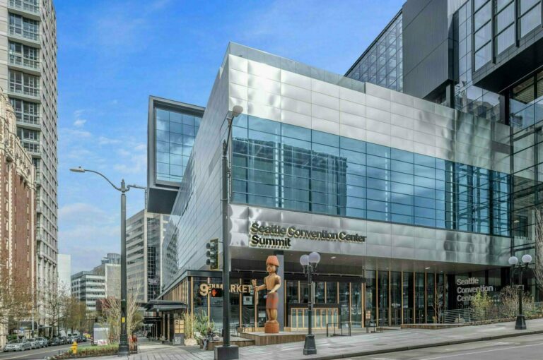 Street view of the Seattle Convention Center Summit building. It features modern glass architecture with reflections and a "Seattle Convention Center Summit" sign. Surrounding buildings are visible under a blue sky.