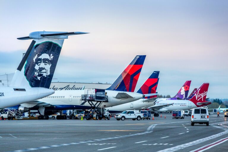 Airplanes from various airlines, including Alaska, Delta, and Virgin America, are parked at an airport terminal at sunset. A white van is on the tarmac, and the sky is a soft gradient of pink and blue.
