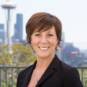 A smiling person with short brown hair is standing outdoors. They are wearing a black blazer and a pink top. The background features a blurred view of the Space Needle and a cityscape.