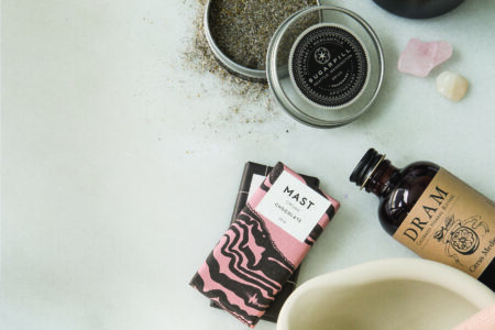 A flat lay of a self-care set on a marble surface, featuring a burning candle, loose tea in a tin, a chocolate bar, a bottle of herbal bitters, wooden incense, and a mortar and pestle. Scattered rose quartz stones complete the arrangement.
