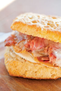 A close-up of a biscuit sandwich with crispy bacon and melted cheese between two halves of a flaky biscuit. The sandwich is placed on a wooden surface, highlighting its golden-brown texture.