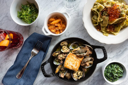 A table setting with a cast iron dish of seafood and bread, a bowl of pasta topped with roe, bowls of greens, a dish of seasoned potatoes, and a glass of red drink with citrus slices. A fork rests on a blue napkin.