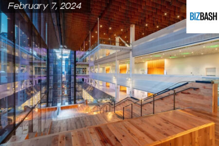 Spacious, modern interior of a convention center with large windows, wooden floors, and multiple levels. Logo: 