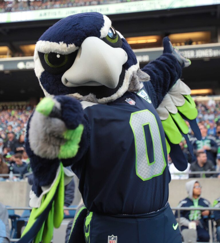 A large, blue and green bird mascot dressed in a sports jersey poses energetically with arms outstretched at a crowded football stadium. The crowd is visible in the background, adding to the lively atmosphere.