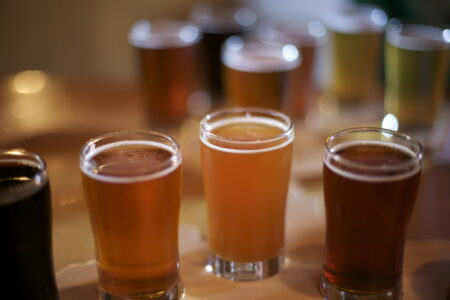 A variety of small glasses filled with different types of beer are arranged on a wooden table. The colors range from light amber to dark brown, suggesting a beer tasting flight. The background is softly blurred.