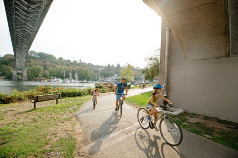 A group of people biking on a path under a large bridge, with a scenic view of water and sailboats in the background. They are wearing helmets, and the sun is shining brightly, casting long shadows on the ground.