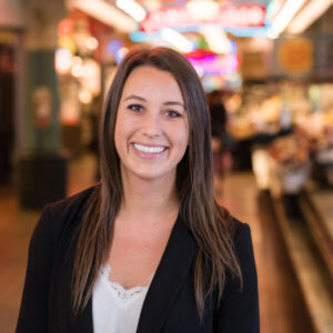 A woman with long brown hair and a white top, wearing a black blazer, smiles at the camera. She stands indoors in a market setting with colorful blurred lights and stalls in the background.