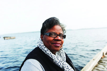 A person wearing glasses and a patterned scarf stands on a dock with a lake in the background. The sky is overcast, and the water is calm.