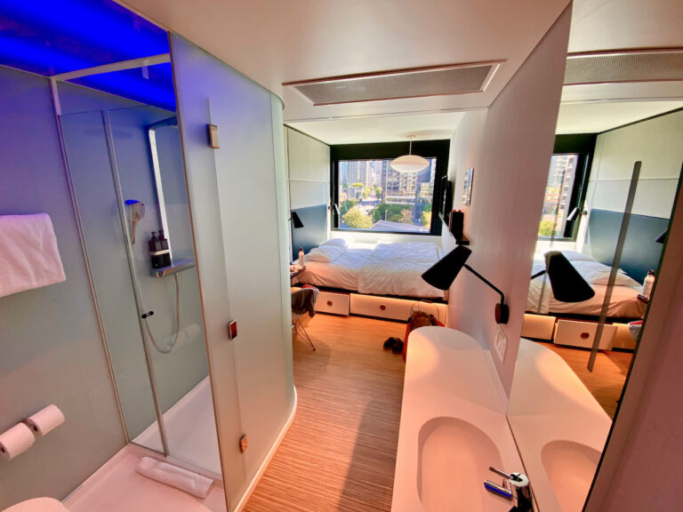 A modern hotel room features a compact glass-enclosed shower and a minimalist bathroom with a double sink. Beyond the partition, there is a cozy bedroom with a neatly made bed, two black wall lamps, and a large window with a city view.