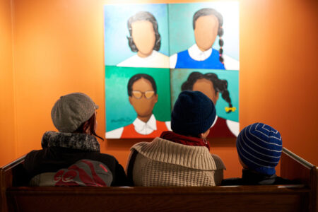 Three people wearing winter clothing sit on a bench facing a painting of six faceless figures on an orange wall. The painting features bright colors and varied hairstyles. The viewers' backs are to the camera.