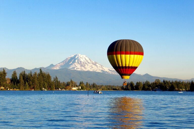 A colorful hot air balloon with red, yellow, and black stripes floats over a serene lake. A distant snow-capped mountain rises in the background under a clear blue sky. Pine trees line the shore, completing the scenic view.