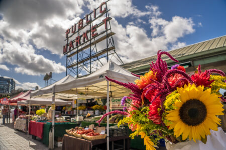 A vibrant bouquet of sunflowers and other colorful flowers in the foreground with a bustling outdoor market scene behind. The iconic 