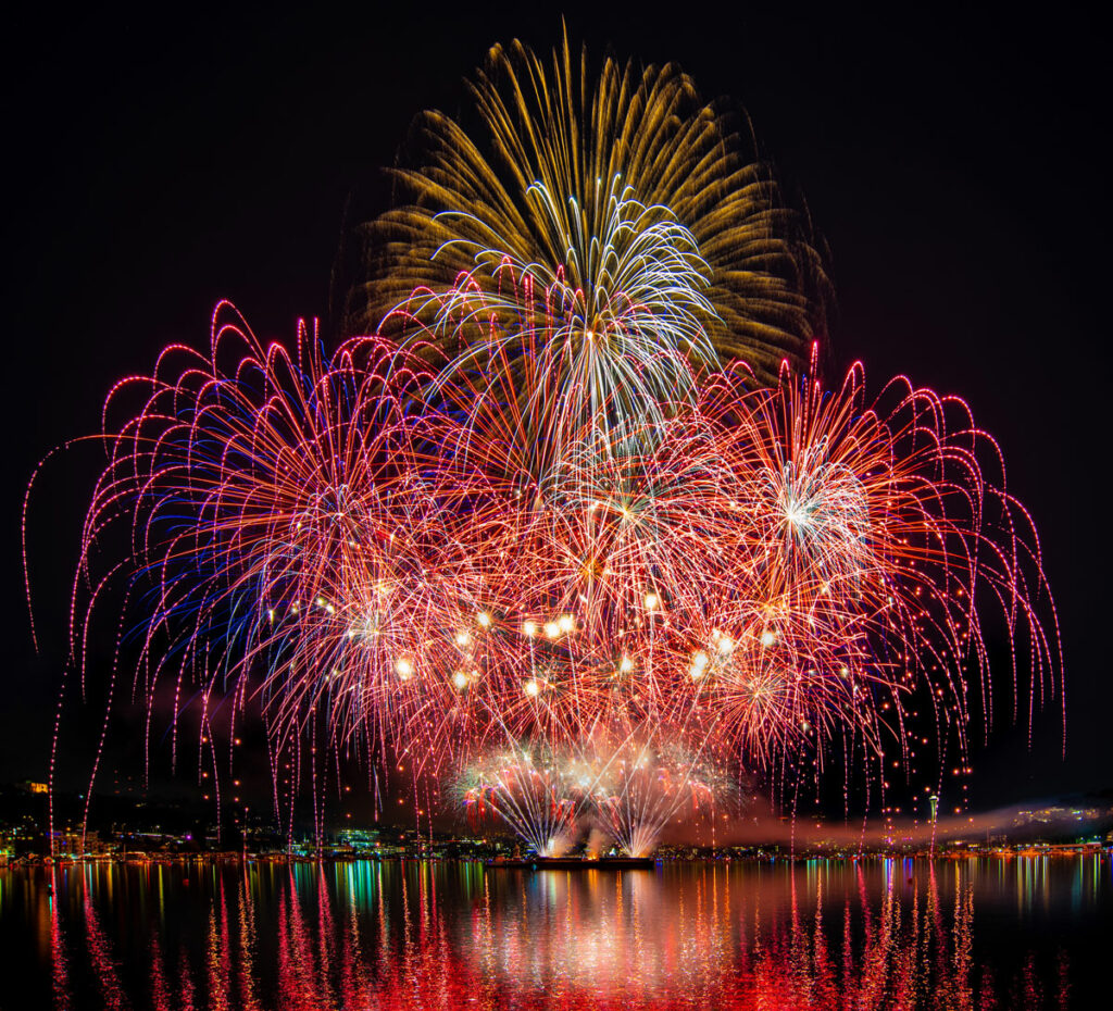 Fireworks illuminate the night sky above the water, creating a mesmerizing display of colors and patterns. The fireworks burst in colors of red, yellow, white, and blue. The colors reflect off the dark water.