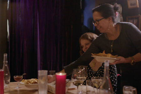 A woman wearing glasses and a dark top places a plate of food on a table surrounded by drinks, including wine and a red candle. She smiles while serving, and another person is partially visible in the background. Dark curtains hang behind them.