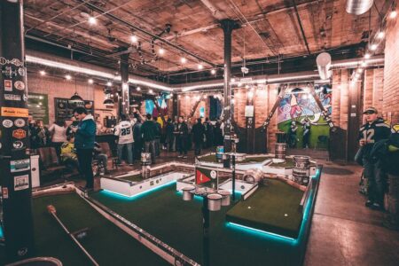A bustling indoor mini-golf course with people gathered around. The room has exposed brick walls, string lights, and a lively atmosphere. Several mini-golf holes with obstacles and neon lights are visible on the green turf.