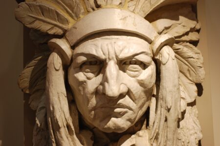 A detailed stone sculpture depicting a stern-looking face wearing a traditional feathered headdress, showcasing intricate carving and shadow contrasts.