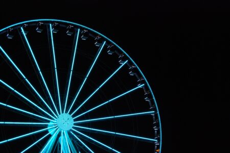 A brightly lit Ferris wheel glows with blue lights against a dark, black sky. Only a portion of the wheel is visible, creating a striking visual contrast.
