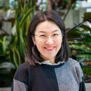 A person with glasses and shoulder-length dark hair smiles at the camera. They are wearing a gray and black sweater. The background features green plants in a blurry indoor setting.