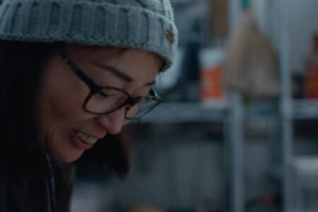 A person wearing glasses and a knitted beanie smiles while looking down. The background is a blurred indoor setting with shelves and various objects.