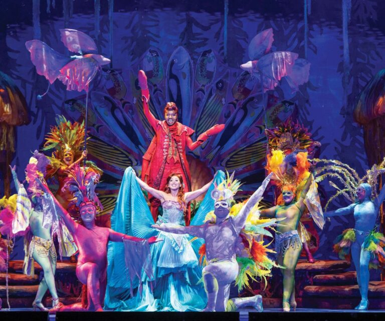 Performers in colorful, elaborate costumes pose on stage. A man in red stands on a raised platform behind a woman in blue. Surrounding them are dancers dressed as vibrant exotic animals, set against a fantastical forest backdrop.