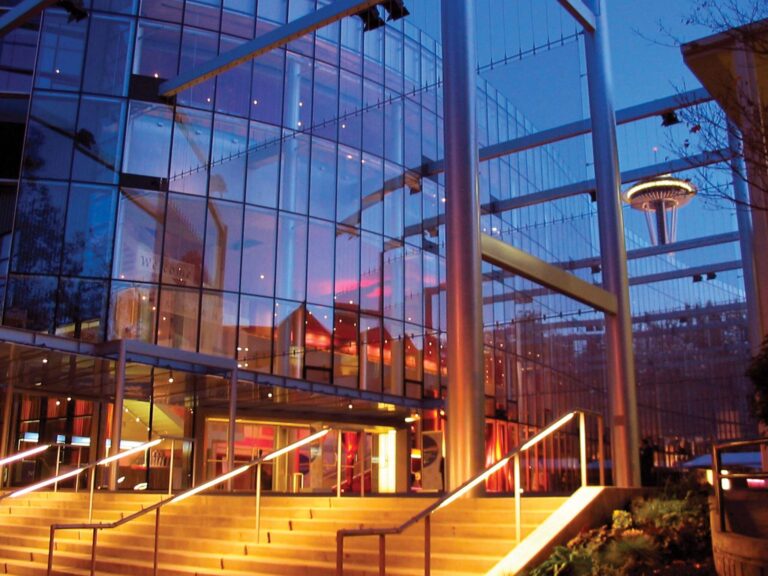 A modern glass building with illuminated stairs at dusk, reflecting the city lights. In the background, the iconic Space Needle is visible against the evening sky, adding to the urban skyline.