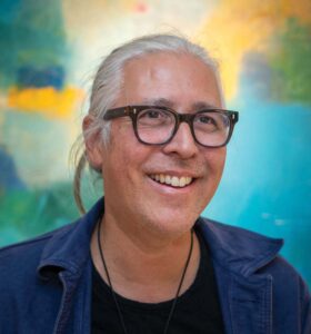 A person with long gray hair and glasses smiles against a colorful abstract background. They are wearing a dark shirt and a blue jacket.