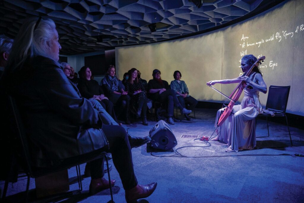 A performer plays the cello in a dimly lit room with an audience seated around. They are wearing a flowing outfit, and the ambiance is artistic with a wavy ceiling and illuminated text on the wall.