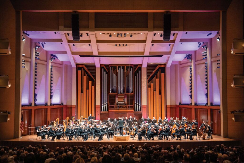 An orchestra performs on a large, well-lit stage with a prominent pipe organ in the background. The musicians are seated, and a conductor stands in front, leading them. Audience members are visible in the foreground.