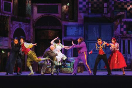A lively theater performance featuring actors in colorful, eclectic costumes. The stage is set with vibrant set pieces and the actors are energetically interacting in dramatic poses, suggesting a comedic or whimsical scene.