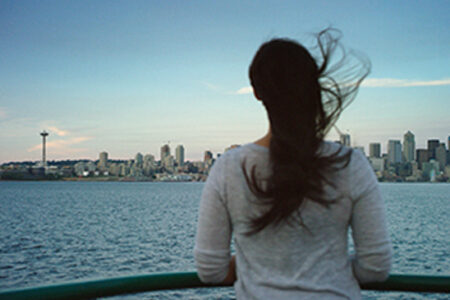 A person with long hair stands on a boat, gazing at a city skyline across the water. The Space Needle is visible on the left, and the sky is clear.