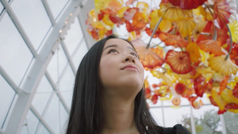 A person with long black hair looks up at vibrant orange and yellow glass sculptures hanging from the ceiling in a glass-enclosed space. The geometric structure and natural light create an open, airy atmosphere.