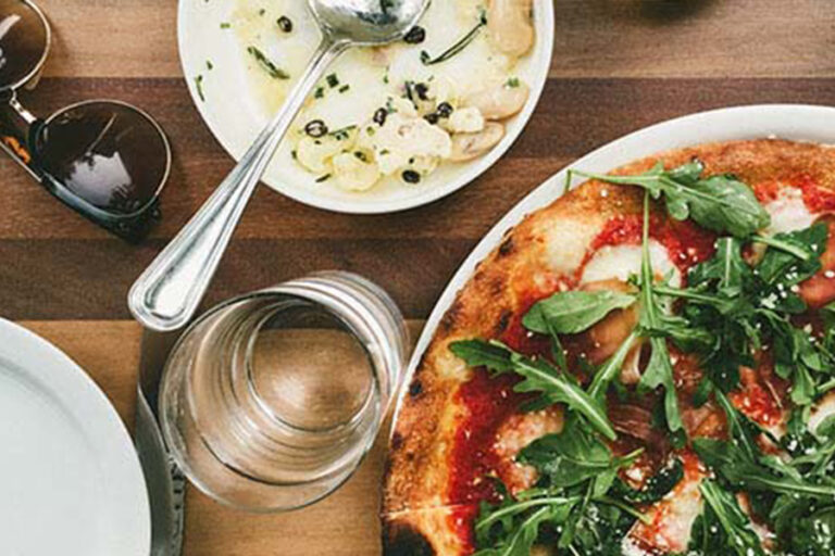 A wooden table with a pizza topped with arugula, a glass of water, a small bowl of pasta salad with a spoon, and a pair of sunglasses.