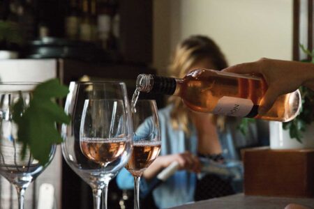 A person is pouring rosé wine into one of three wine glasses on a counter. In the background, another person sits out of focus, surrounded by shelves filled with bottles. The scene has a cozy, relaxed atmosphere.