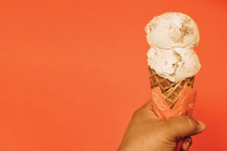 A brown hand holding a waffle cone wrapped in orange paper with two scoops of white ice cream against an orange backdrop