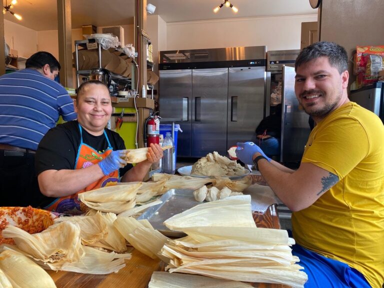Two people smile at the camera as they sit at a workstation, creating handmade tamales from scratch.