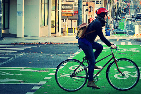 A cyclist wearing a helmet and backpack rides a bike on a bright green bike lane in an urban setting. The road has markings for bikes, and there's a parking sign and buildings in the background. The street is lined with fall foliage.