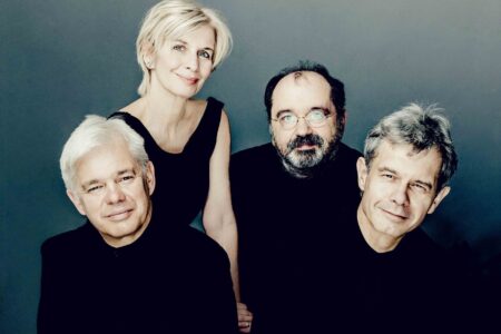 Four people posing against a gray background. Three men and one woman are dressed in black. Two men are seated on stools, while the other two are standing behind them, smiling softly.