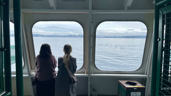 Two people stand on a ferry, gazing out of large windows at a calm body of water. The distant city skyline is visible under a cloudy sky. A trash bin is in the foreground.