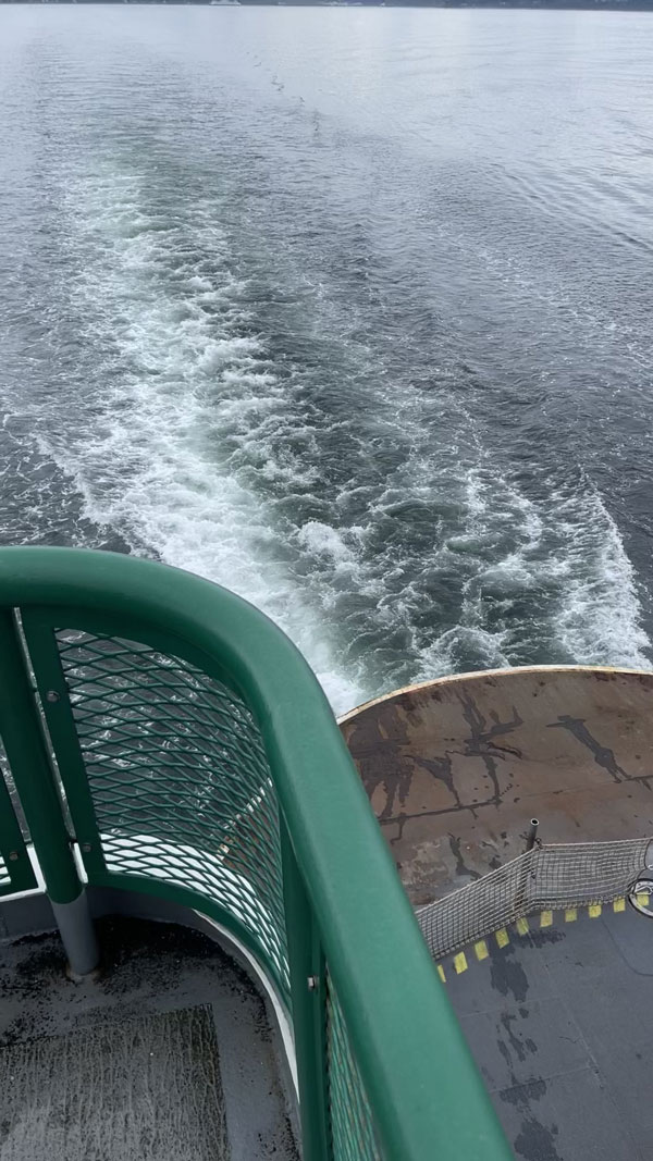 View from a ferry deck showing the wake left in the water as the vessel moves forward. The railing is painted green, and the deck has a weathered appearance with some yellow markings. The surrounding water is calm and expansive.
