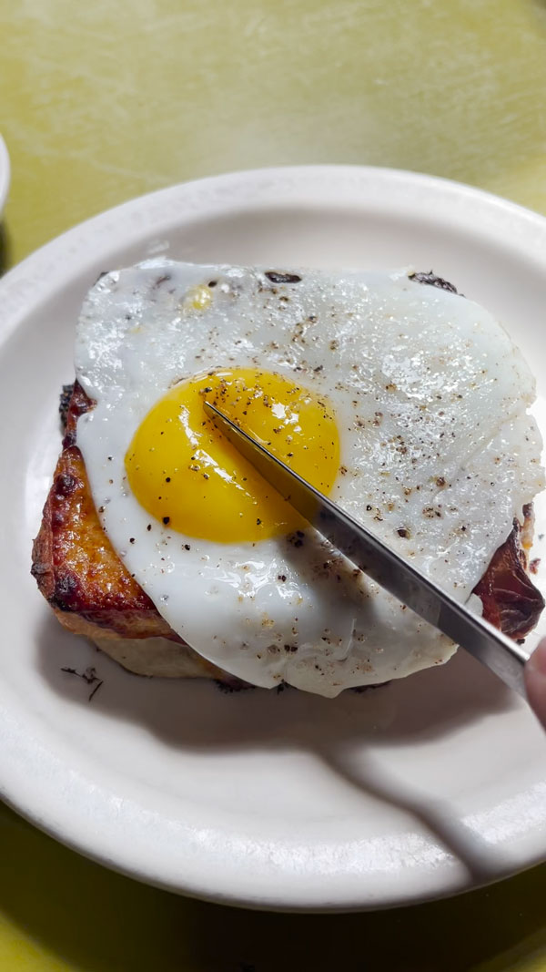 A knife cuts into a sunny-side-up egg with a runny yolk, placed on top of a piece of toast. The toast appears to have melted cheese and tomato sauce on it, and is served on a white plate.