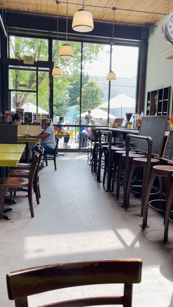 Interior of a café with large windows letting in natural light. People sit at tables with laptops and chairs. Outside, more people are visible under umbrellas. The café has wooden furniture and a casual atmosphere.