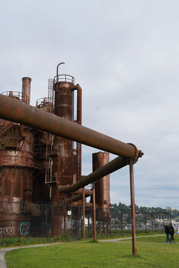 An image of Gas Works Park in Seattle, featuring rusted industrial structures against a cloudy sky. A grassy field surrounds the machinery, with walking paths and distant views of a water body and urban skyline. A few people are in the distance.