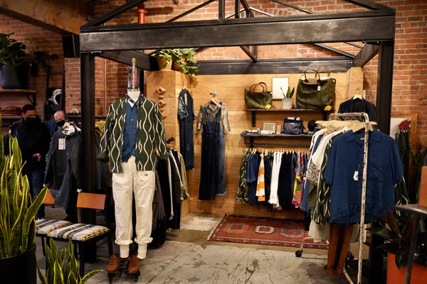 A trendy clothing store interior with a mannequin dressed in a patterned shirt and white pants. Racks of clothes, bags, and plants decorate the space, which features brick walls and a cozy, industrial vibe.
