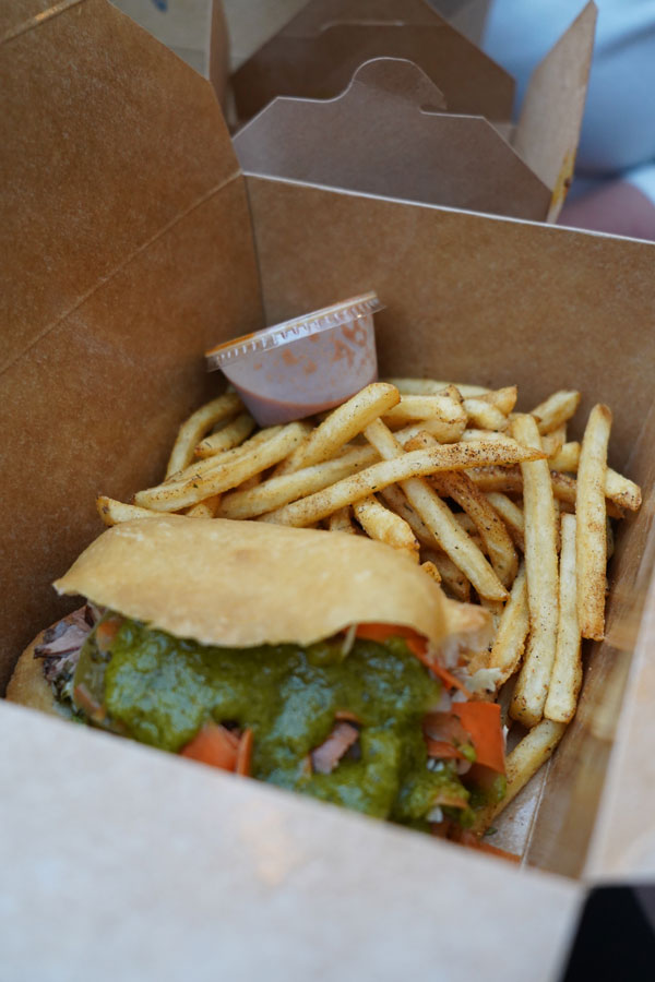 A takeout box containing a sandwich topped with green sauce and sliced tomatoes, accompanied by a side of seasoned french fries. A small container of sauce is placed next to the fries.